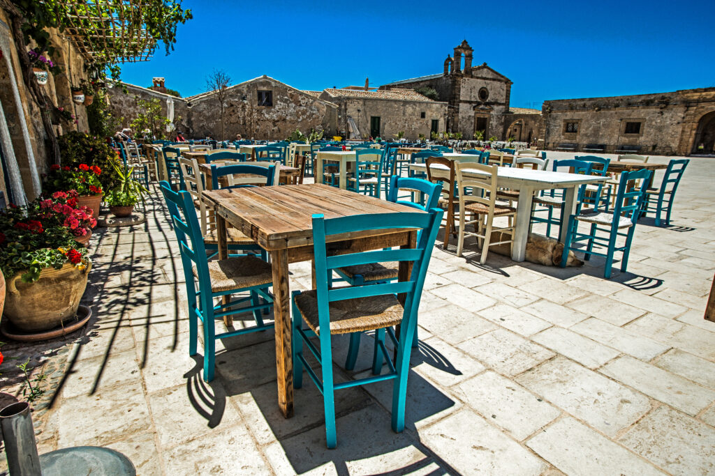 Blue and white chairs and outdoor tables in Piazza Regina Margherita in Marzamemi, with a dog resting beneath a table away from the heat of Italy.