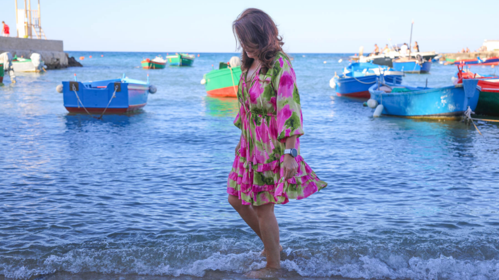 Michelle Tissi walking along the water among colorful fishing boats in Marzamemi, Sicily
