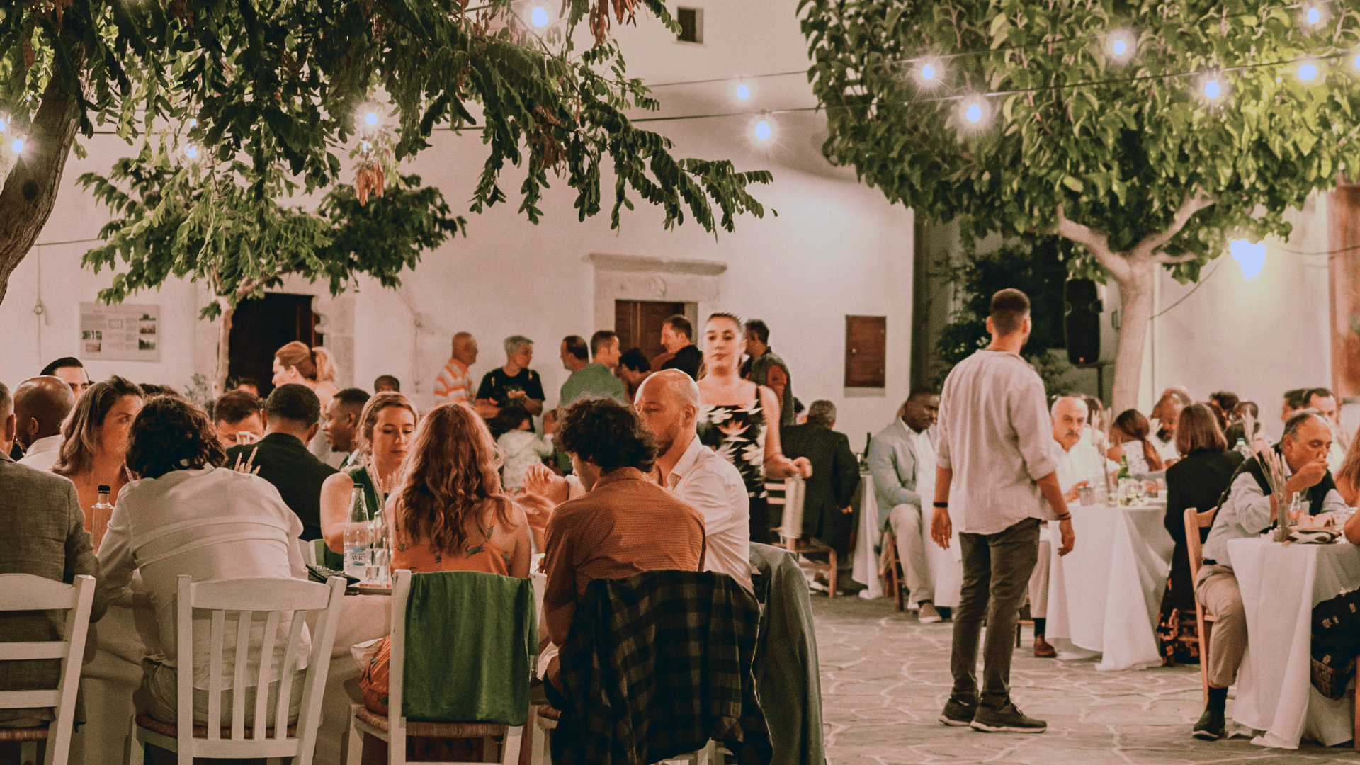 Guests dining at outdoor tables beneath leafy trees and string lights in a courtyard.