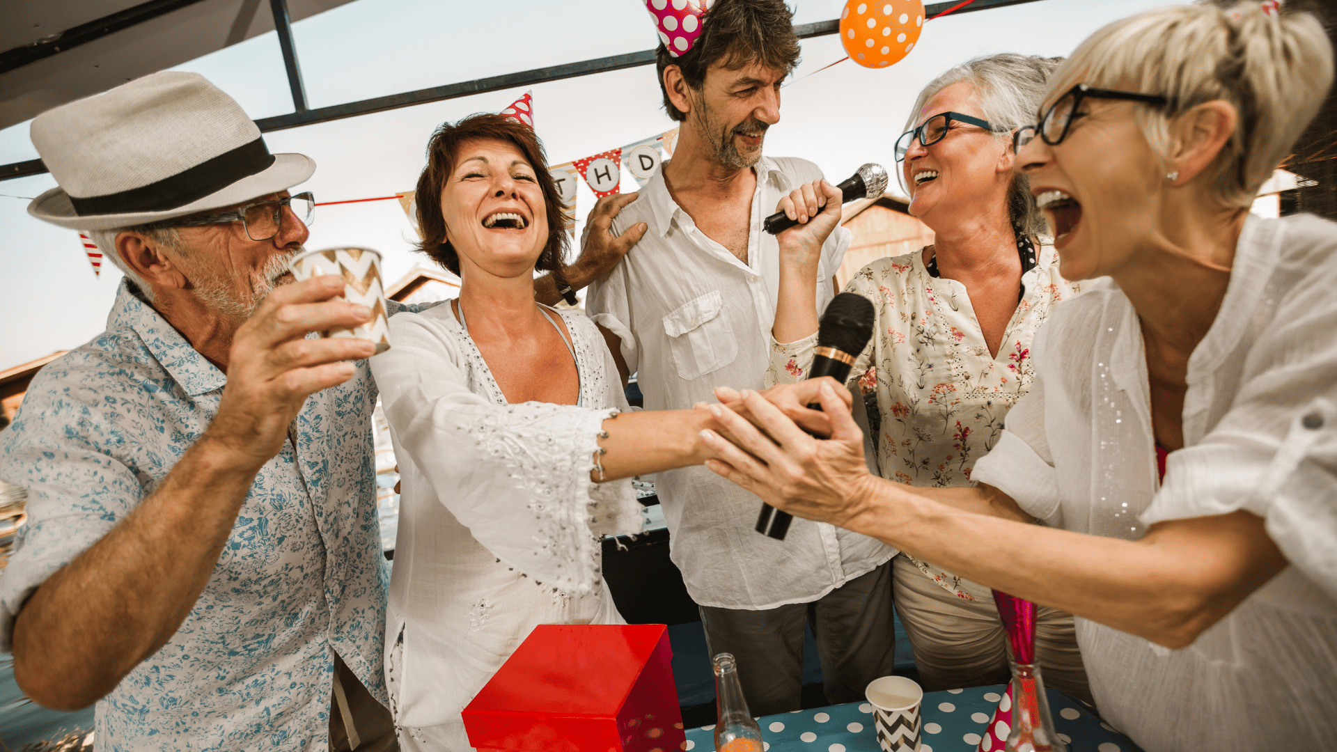 Five adults laughing and singing into microphones at a birthday party, with drinks and party decorations.