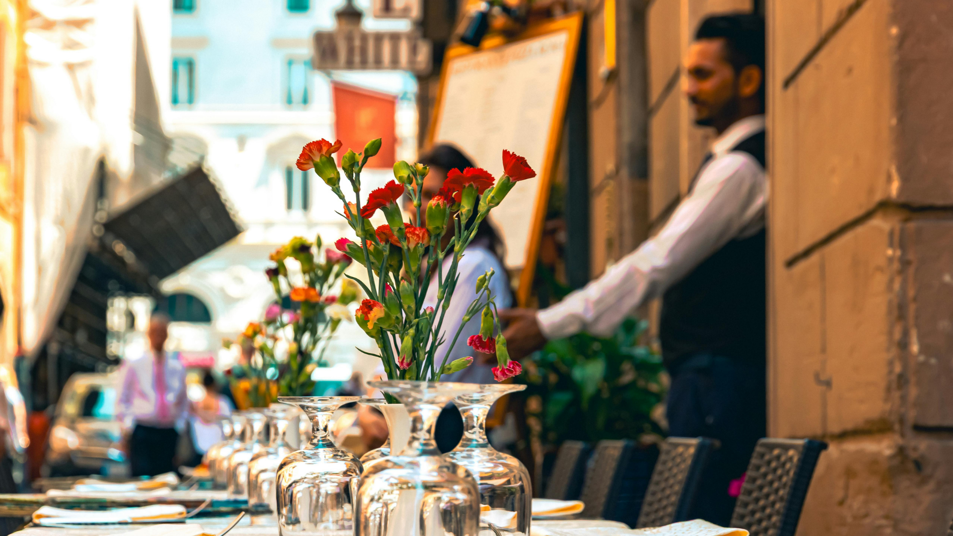 Outdoor restaurant tables set with plates, inverted wine glasses and red flowers, with a waiter in the background.