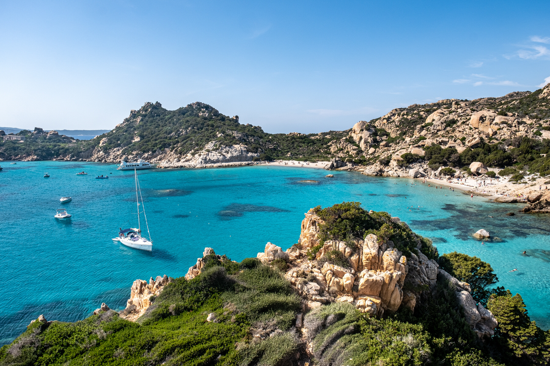 Boats in bright blue water beneath the rocky coastline of Isola di Spargi in Sardinia.